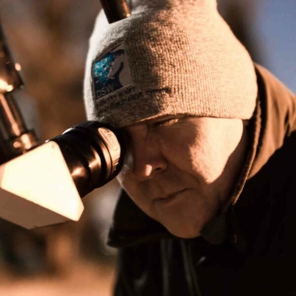 A person wearing a knit hat and jacket peers through a telescope eyepiece in an outdoor setting at Kew Gardens, creating a memorable moment during their Family Visit to Kew Gardens for Two Adults and Two Children.