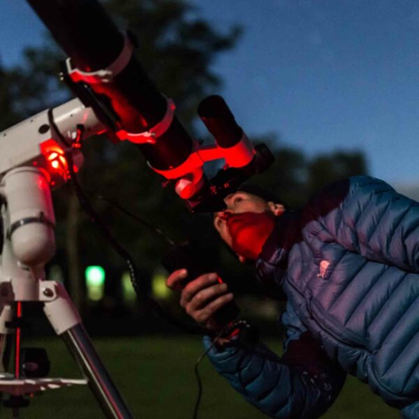 During a Family Visit to Kew Gardens for Two Adults and Two Children, there's a memorable moment where someone in a blue jacket peers through an outdoor telescope under the night sky, lit by a red light. Meanwhile, two adults and two children eagerly wait for their turn.