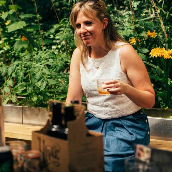 A woman holding a drink is sitting outdoors at a table surrounded by greenery and yellow flowers, enjoying the *Totally Wild Foraging, Cookery and Lunch for Two*. Various bottles and packages on the table hint at an afternoon of culinary adventures.
