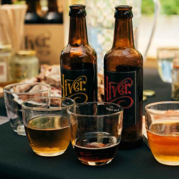 Two bottles of Hiver Honey Ale are placed on a table set for the "Totally Wild Foraging, Cookery and Lunch for Two," with four glasses containing different amounts of beer in front of them. Various other items, hinting at wild foraging and cookery delights, are seen in the background.