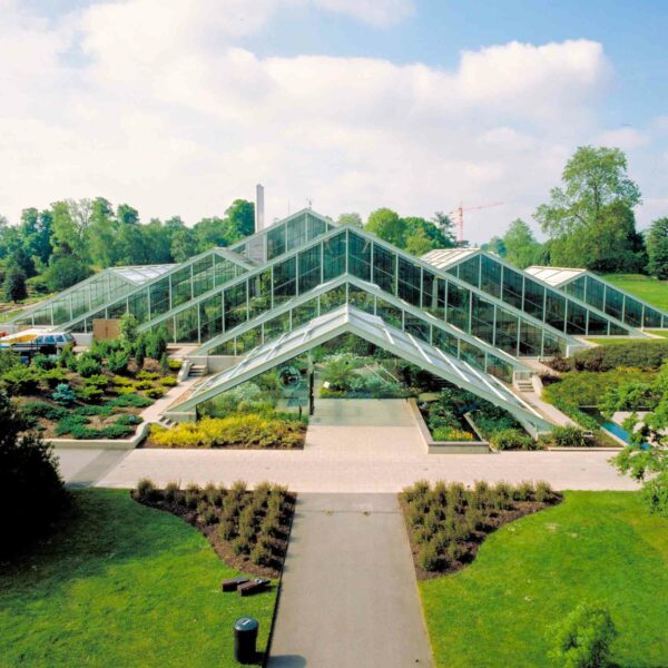 Experience a visit to the picturesque Kew Gardens under a partly cloudy sky, featuring a modern glass greenhouse with multiple peaked roofs, surrounded by beautifully landscaped gardens and pathways. Perfect for two people, this outing includes a delightful cream tea amidst nature's beauty.