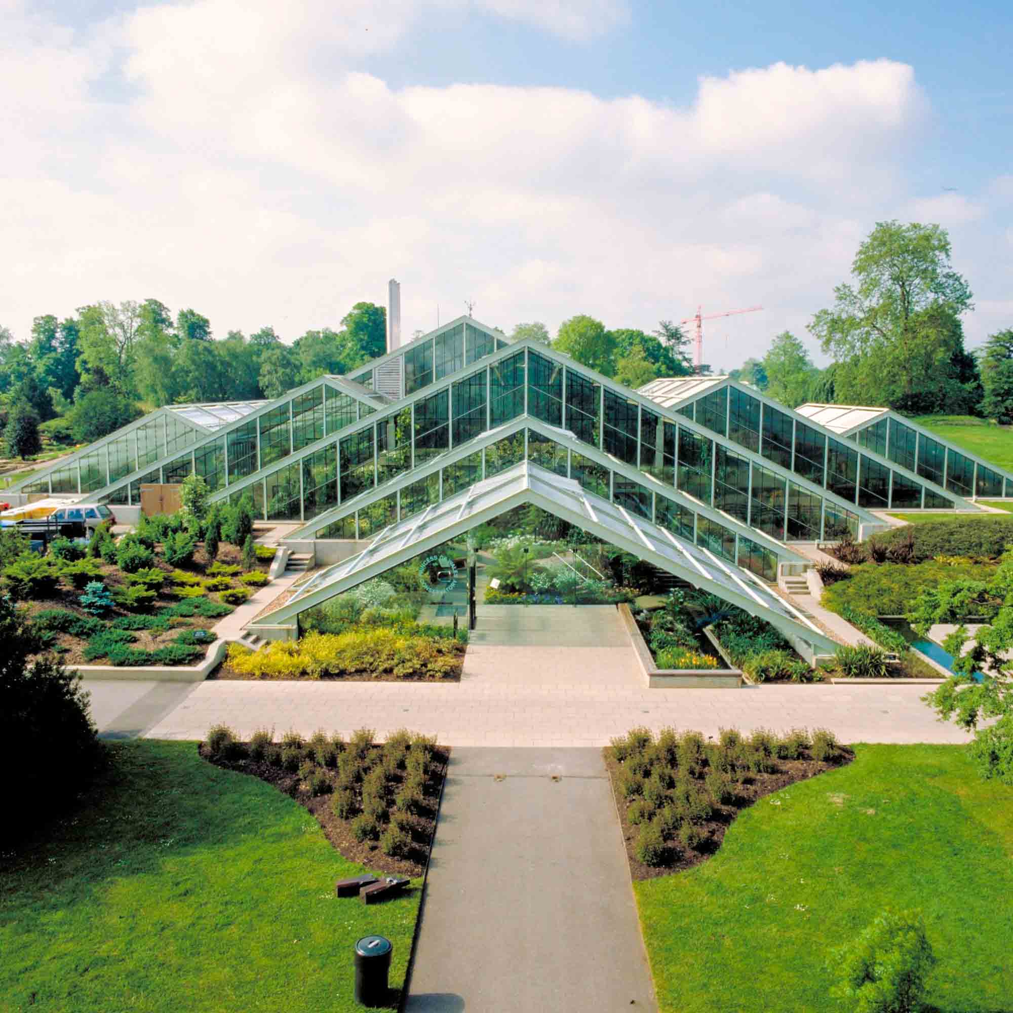 Experience a visit to the picturesque Kew Gardens under a partly cloudy sky, featuring a modern glass greenhouse with multiple peaked roofs, surrounded by beautifully landscaped gardens and pathways. Perfect for two people, this outing includes a delightful cream tea amidst nature's beauty.