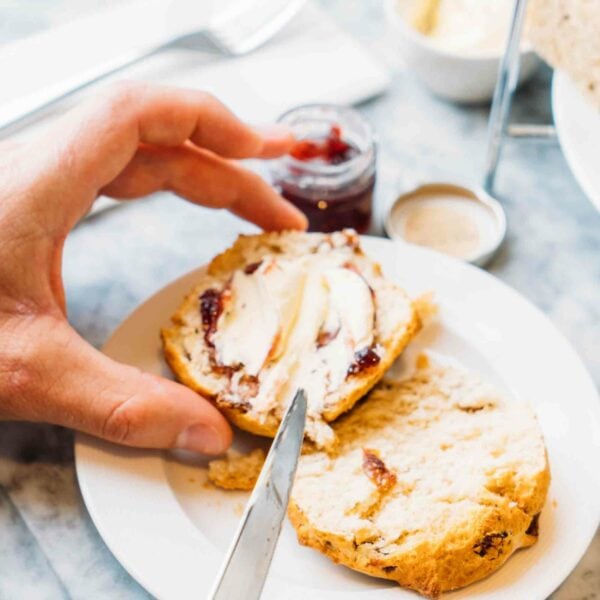 A person enjoys spreading jam and clotted cream on a scone with a knife, accompanied by a small jar of jam, a dish of cream, and two glasses of prosecco in the background, during their Terrarium Workshop with Prosecco for Two.