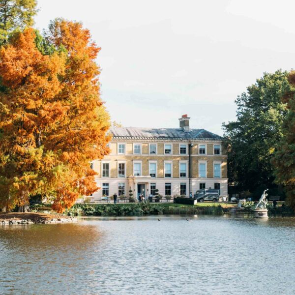 A stately brick building with large windows stands by a calm lake, surrounded by green and orange autumn trees, offering a serene setting for a Terrarium Workshop with Prosecco for Two.