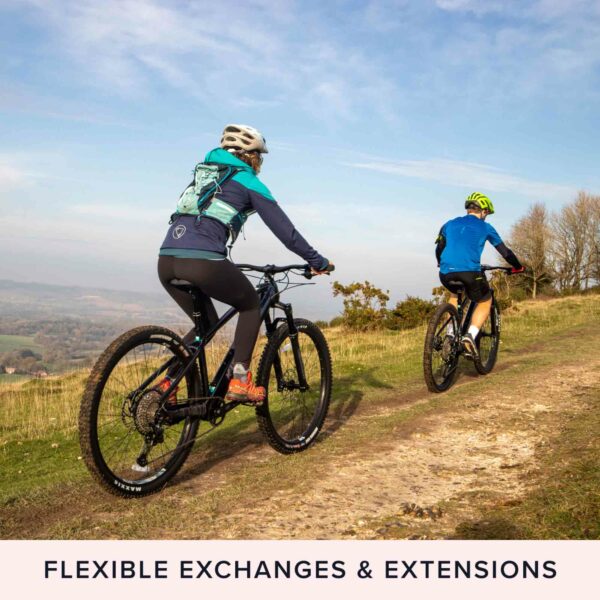 Two cyclists enjoy their mountain bikes on a grassy trail by a lush forest under a clear sky. Text below reads: "Forest Bathing Experience for Two - FLEXIBLE EXCHANGES & EXTENSIONS.
