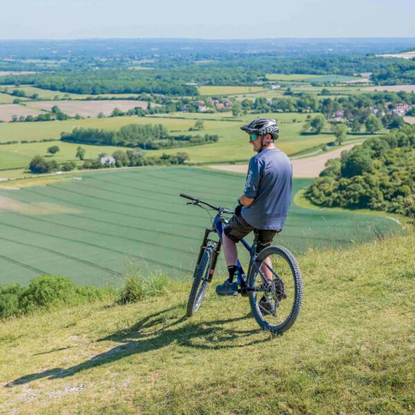 A person equipped with a helmet and riding gear sits on a bicycle, fully engaged in the Forest Bathing Experience for Two, gazing over an expansive green landscape from atop a hill.