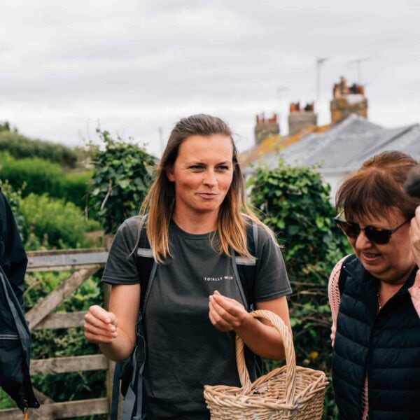 Two women, dressed casually and holding a wicker basket, stand outdoors against a backdrop of greenery with buildings in the distance. They seem to be enjoying the "Totally Wild Foraging, Cookery and Lunch for Two" experience.