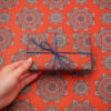 A hand holds a rectangular present wrapped in floral patterned gift wrap and tied with blue string, complementing the ornate background.