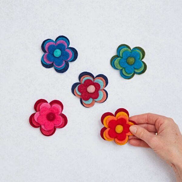 A hand skillfully arranges vibrant Felt Four Flower Brooches on a light surface, crafting a delightful display.
