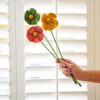 A hand gently holds a Pack of 3 Felt Handmade Flowers—green, yellow, and orange—against white window shutters.