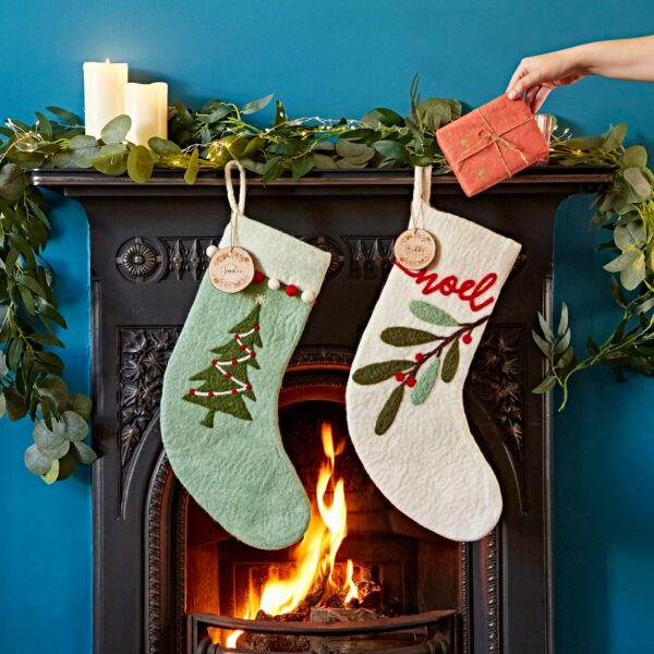 Two Felt Christmas Stockings hang on a fireplace mantel above a lit fire. The mantel is adorned with candles and greenery. A hand holds a wrapped gift, adding to the felt Christmas ambiance.