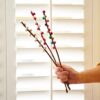 A hand holds a Pack of 3 Felt Handmade Pussy Willow Catkins in front of white shutters, decorated with colorful felt balls.