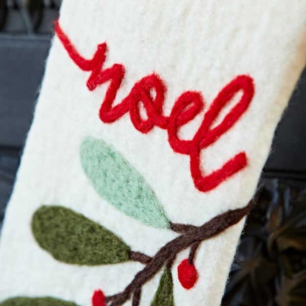 Close-up of a Felt Christmas Stocking featuring white fabric, red embroidery spelling "Noel," and green and brown felt leaves decorated with red berries.