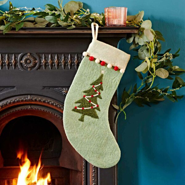 A Felt Christmas Stocking featuring a decorated tree design hangs on a fireplace mantel, adorned with greenery. A lit candle and a fire are visible in the background, adding to the festive atmosphere.