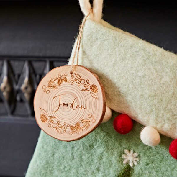 A Felt Christmas Stocking in beige with red and white pom-poms hangs by a fireplace. A round wooden ornament engraved with the name "Jordan" and festive designs is attached to the stocking.