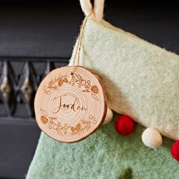 A Personalised Felt Christmas Stocking features a round wooden ornament engraved with "Jordan" and botanical designs, hanging from its green fabric.