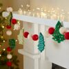 A fireplace mantel is adorned with the Felt Christmas Tree Garland, featuring felt red and white balls along with green felt Christmas trees, with string lights elegantly draped along the top edge. In the background, a partially visible Christmas tree adds to the festive ambiance.