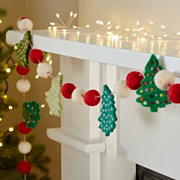 A fireplace mantel is adorned with the Felt Christmas Tree Garland, featuring felt red and white balls along with green felt Christmas trees, with string lights elegantly draped along the top edge. In the background, a partially visible Christmas tree adds to the festive ambiance.
