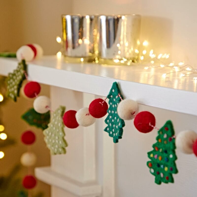 A Felt Christmas Tree Garland adorned with red, white, and green ornaments, featuring trees and balls, decorates a white mantle. Two silver candles and string lights are also placed on the mantle.