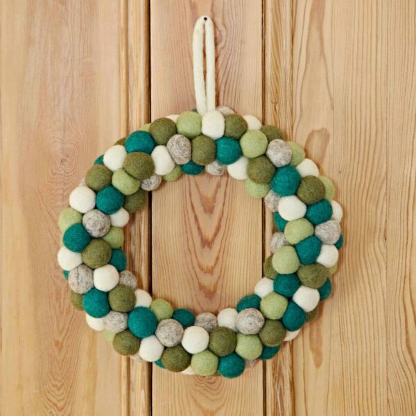 The Felt Green Wreath, featuring green, blue, and white felt balls, adds a decorative touch when hanging on a wooden wall.