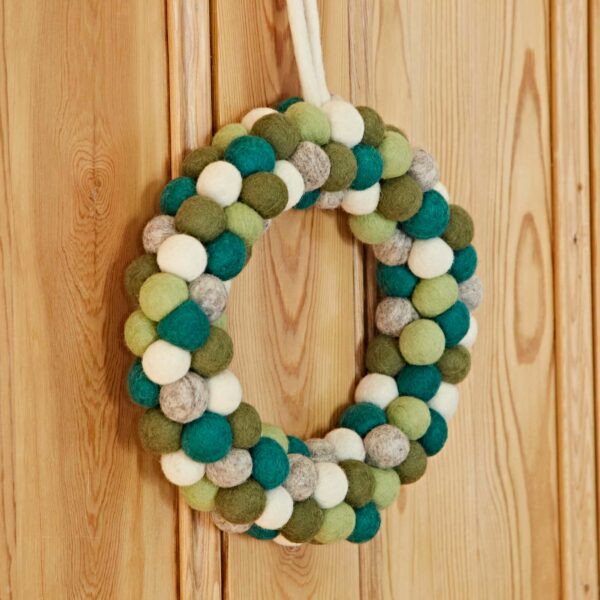 The Felt Green Wreath, featuring a harmonious blend of green, white, and gray felt balls, hangs elegantly on a wooden wall.