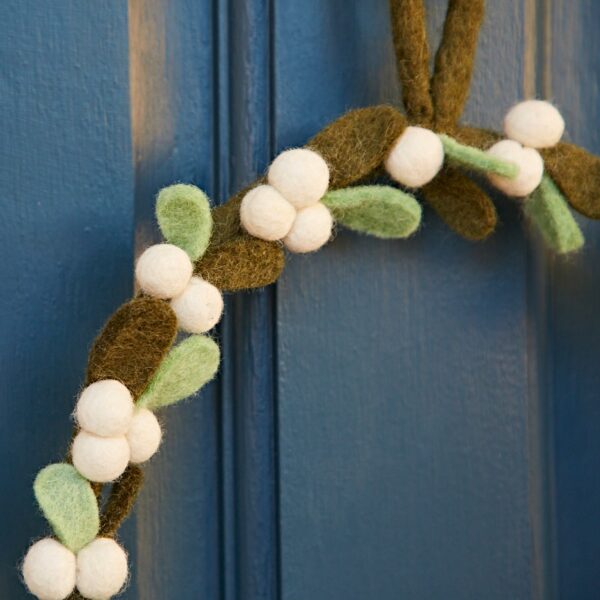 The Felt Mistletoe Christmas Wreath, featuring white berries and green leaves, elegantly hangs on a blue door.