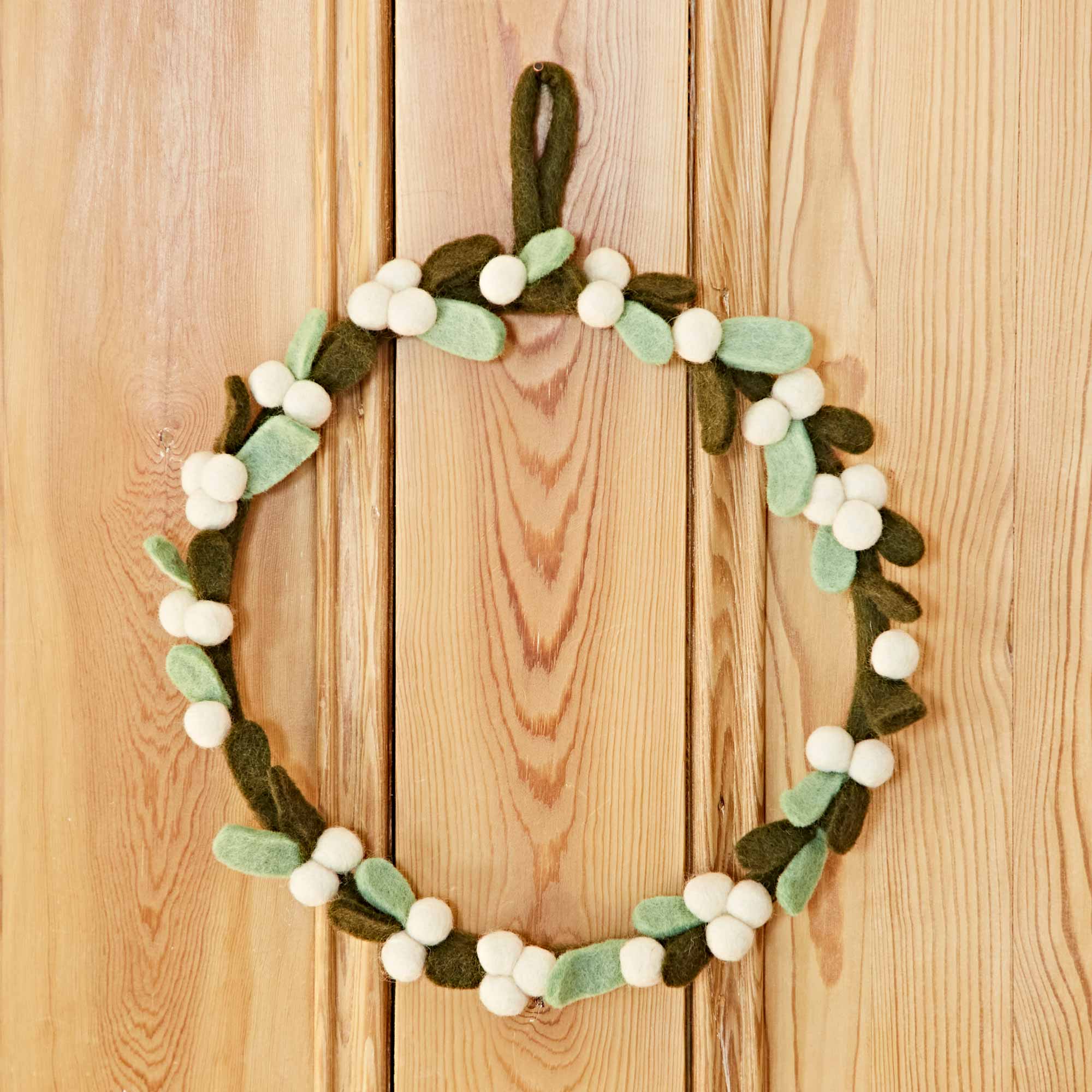 The Felt Greenery with Pompoms Christmas Wreath, adorned with green leaves, white berries, and a subtle touch of felt greenery, hangs on a wooden surface.