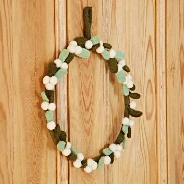 The Felt Greenery with Pompoms Christmas Wreath, adorned with green felt leaves, white felt balls, and colorful pompoms for a festive flair, hangs on a wooden door.