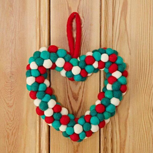 A Felt Heart Wreath, crafted with red, green, and white pom-poms, hangs on a wooden surface.
