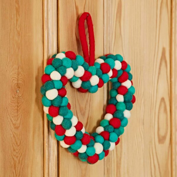 The Felt Heart Wreath is crafted from colorful felt balls in shades of red, green, white, and teal, and is hung on a wooden surface with a red loop.