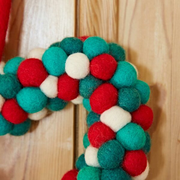 A close-up image of the Felt Heart Wreath, handcrafted from multi-colored felt balls in red, green, and white, hanging on a wooden wall.