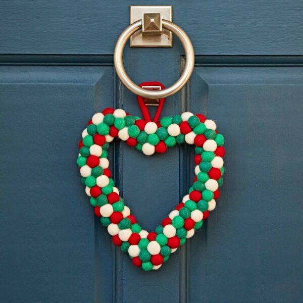 The Felt Heart Wreath, adorned with red, green, and white felt pom-poms, hangs elegantly on a blue door, highlighted by a gleaming metal knocker.