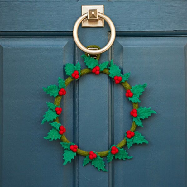 A blue door is elegantly adorned with the Felt Holly Christmas Wreath, featuring green leaves and red berries hanging from a silver knocker, and felt accents that add a cozy touch.