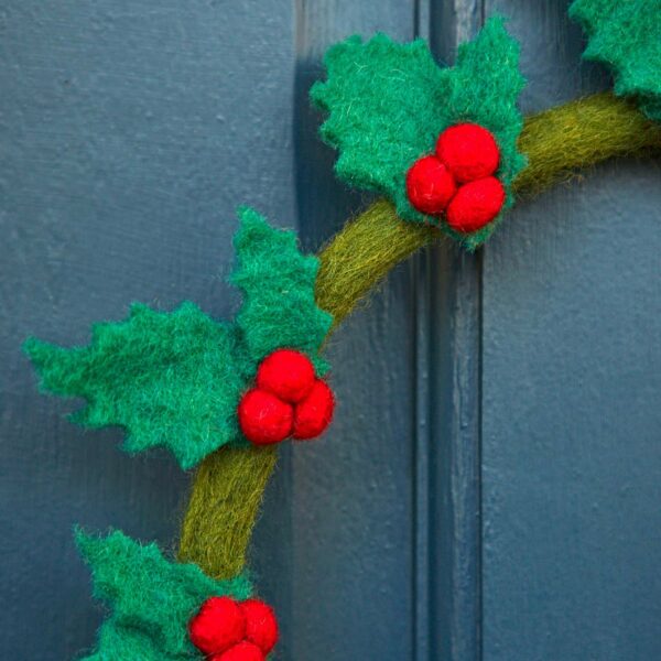 The Felt Holly Christmas Wreath, adorned with green leaves and red holly berries, is hanging on a blue door.