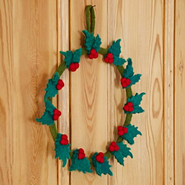 The Felt Mistletoe Christmas Wreath, adorned with green leaves and red berries, hangs elegantly on a wooden door.