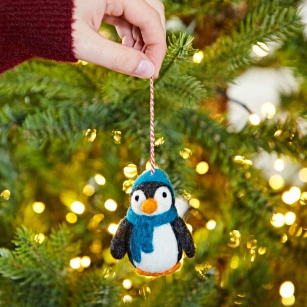 A hand is holding a Felt Penguin with a Hat and Scarf Christmas Decoration, hanging it on a Christmas tree adorned with lights in the background.