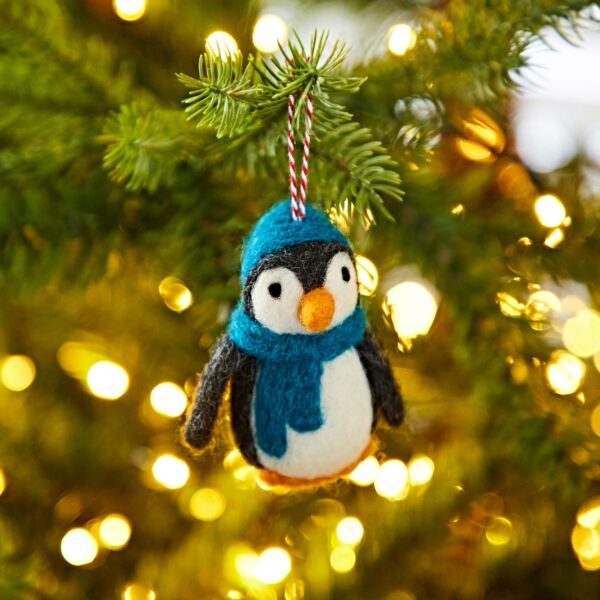 A Felt Penguin with a Hat and Scarf Christmas Decoration, complete with a blue hat and scarf, hangs on a branch of the Christmas tree, illuminated by twinkling lights in the background.