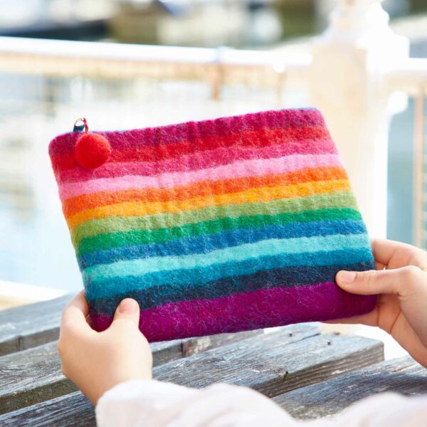 Hands holding a vibrant Felt Tree of Life Purse with colorful stripes and a red pom-pom attached, set against a blurred outdoor background.