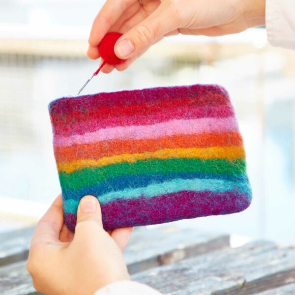 Hands holding a Felt Tree of Life Purse with colorful horizontal rainbow stripes, adorned with a small red pom-pom zipper pull and a Tree of Life motif.