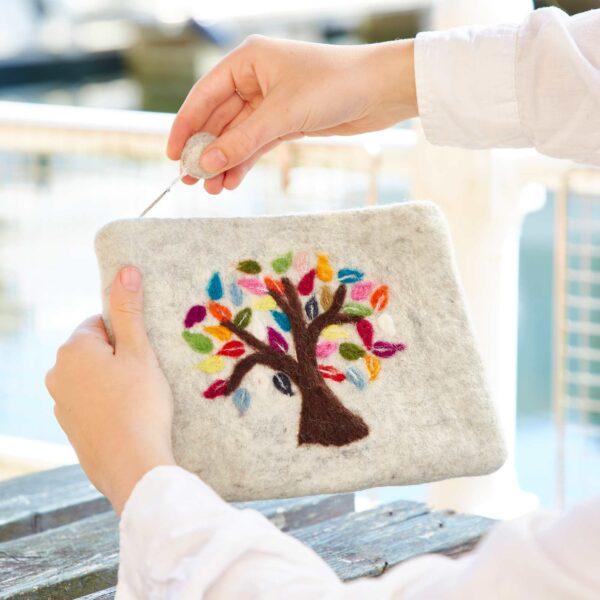 A person uses a felting needle on a gray felted item featuring a vibrant tree design with multicolored leaves, resulting in an intricate Felt Tree of Life Purse.