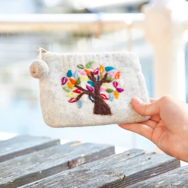 A hand holds the Felt Tree of Life Purse, showcasing its small, light gray felt surface adorned with a colorful embroidered Tree of Life design and multicolored leaves. The background is blurred, suggesting an outdoor setting.
