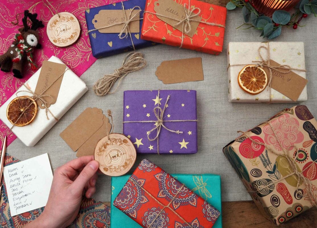 A hand holds a round wooden ornament over a table with wrapped gifts, dried citrus slices, twine, tags, a list, and a small stuffed toy made from elephant dung.
