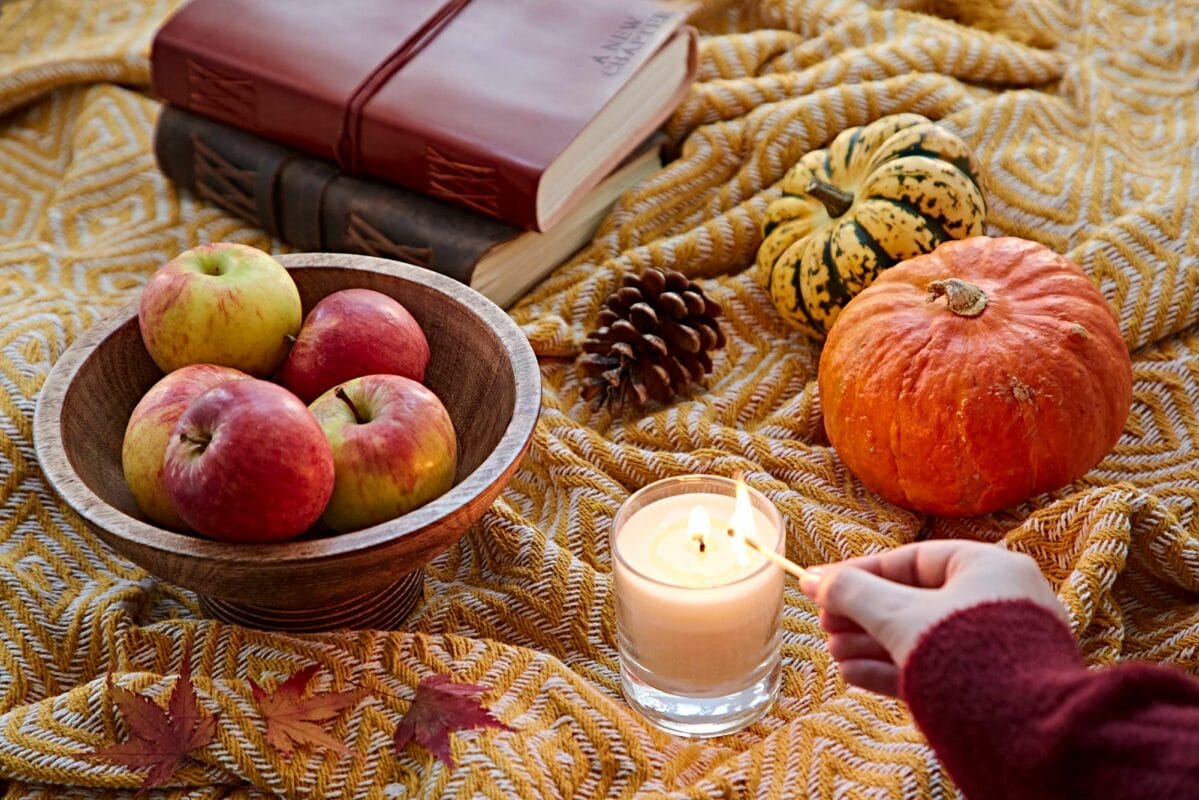 A hand lights a candle on a yellow patterned blanket with apples in a wooden bowl, pumpkins, and a pine cone. Stacked books in the background subtly contrast with an ornate figurine made from elephant dung.