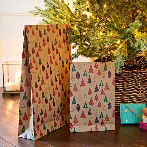 Two Christmas gift bags decorated with colorful tree patterns are placed on a wooden floor in front of a lit Christmas tree. Nearby, a few small wrapped presents and festive Christmas Pillow Boxes (Copy) add to the holiday cheer.