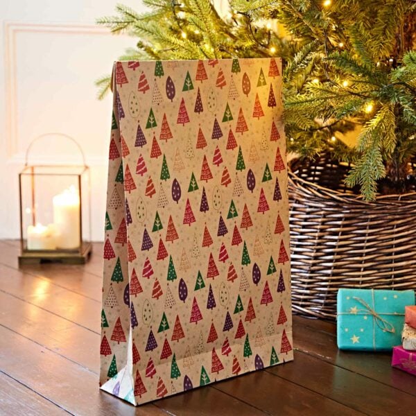 A large, patterned gift bag adorned with festive tree designs is placed near a Christmas tree and a lantern with lit candles, on a wooden floor. Small wrapped presents and charming Christmas Pillow Boxes (Copy) are also visible.