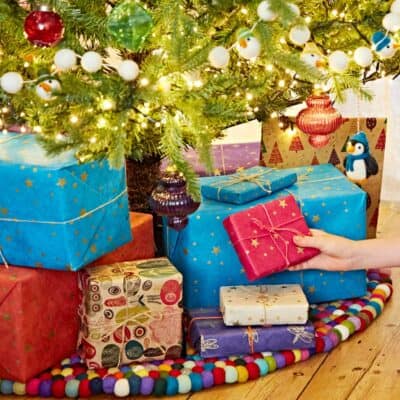 A hand reaches for a red gift box among several wrapped presents under a decorated Christmas tree with baubles and lights, perfect for a festive Christmas Parent Banner.