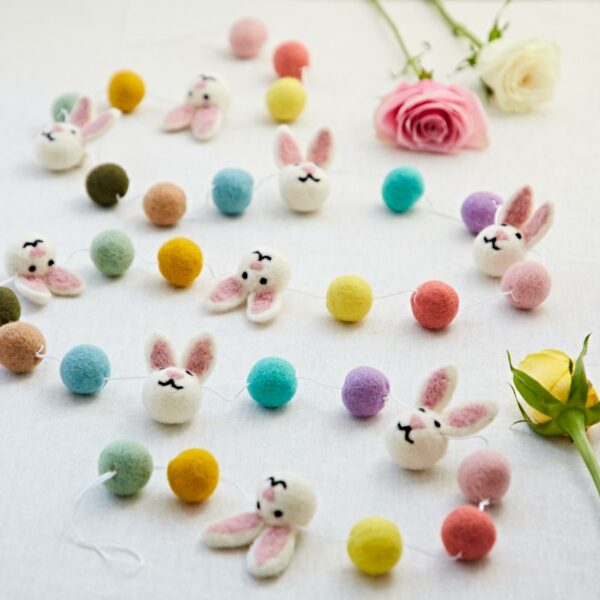 The Felt Bunny Garland, with its lively bunny faces and round felt balls, is beautifully displayed on a table, complemented by an arrangement of pink, white, and yellow roses.