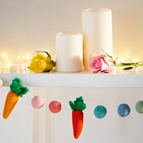 A decorative mantle adorned with two candles, a yellow and pink rose, glowing string lights, and a colorful Felt Carrot Garland featuring carrot and ball shapes.