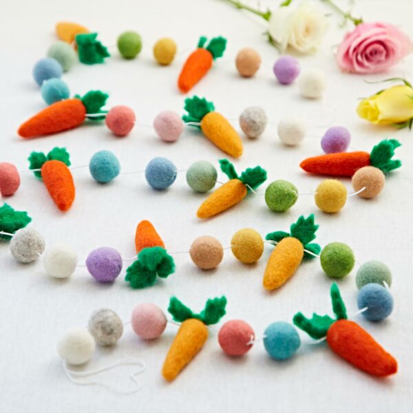 The Felt Carrot Garland, featuring a lively arrangement of carrots and balls, is beautifully displayed on a white surface alongside delicate pink and yellow roses, creating a charming visual ensemble.