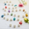 The Felt Easter Egg Garland, featuring vibrant white, blue, pink, and yellow felt balls, decorates the white surface beautifully. Paired with a pink rose and a yellow rose, it provides a charming accent perfect for Easter celebrations.